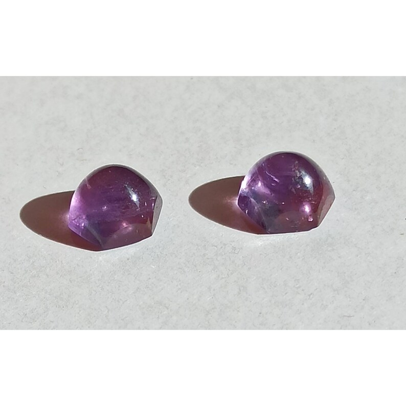 May include: Two faceted, dome-shaped amethyst gemstones. The gems are a deep purple colour with lighter purple and white inclusions. The stones are on a white surface, casting shadows.