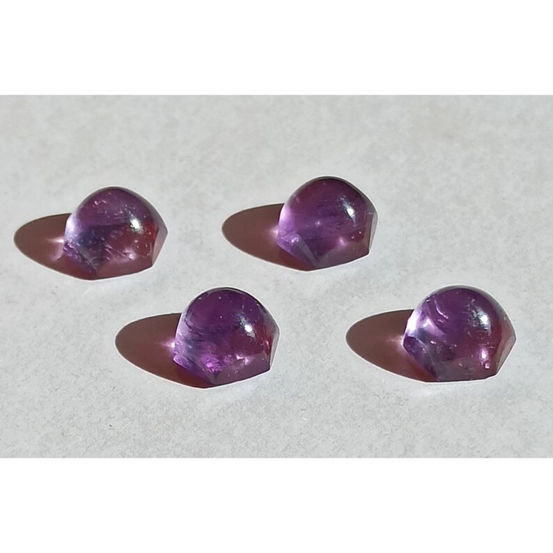 May include: Four faceted, dome-shaped amethyst gemstones. The gems are a deep purple colour with internal swirls and inclusions. Each stone casts a shadow on the white surface.