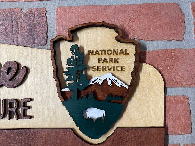 May include: A wooden sign with the words "Welcome to Nature" and a green, brown, and white National Park Service logo. The logo features a mountain, a tree, and a bison.