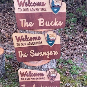 May include: Three wooden signs with a welcome message, the National Park Service logo, and a family name. The signs are stacked on top of each other, with the top sign reading "Welcome to our adventure The Bucks", the middle sign reading "Welcome to our adventure The Swangers", and the bottom sign reading "Welcome to our adventure The Andersons".