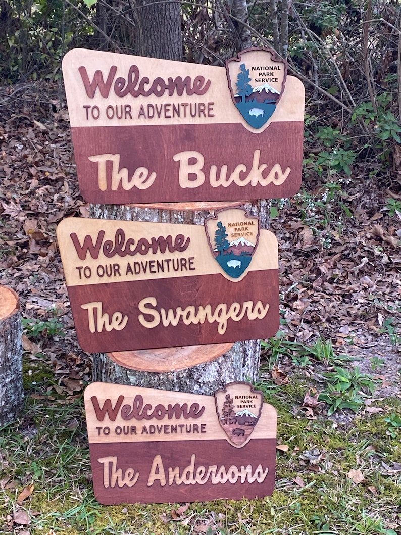 May include: Three wooden signs with a welcome message, the National Park Service logo, and a family name. The signs are stacked on top of each other, with the top sign reading "Welcome to our adventure The Bucks", the middle sign reading "Welcome to our adventure The Swangers", and the bottom sign reading "Welcome to our adventure The Andersons".