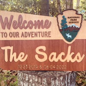 May include: A wooden sign with the text "Welcome to our adventure The Sack 12.23.2019 & 06.04.2022" and a National Park Service logo with a moose.