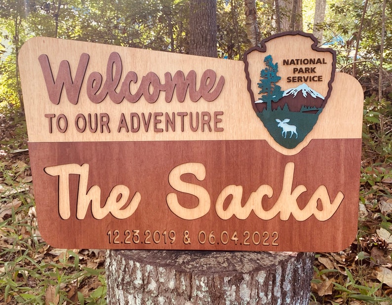May include: A wooden sign with the text "Welcome to our adventure The Sack 12.23.2019 & 06.04.2022" and a National Park Service logo with a moose.