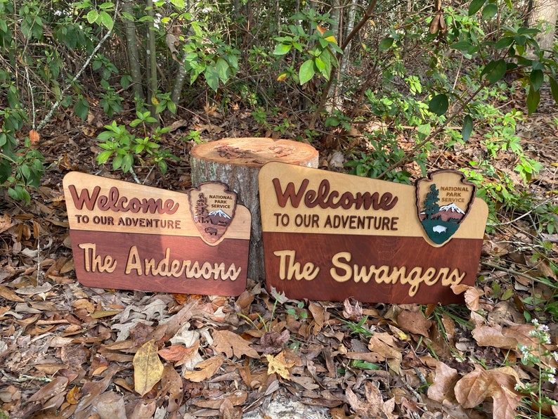 May include: Two wooden welcome signs with the text "Welcome to our adventure" and the family names "The Andersons" and "The Swangers". Each sign features a National Park Service logo with a mountain scene.