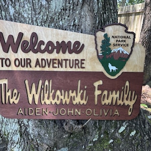 May include: Welcome to our adventure sign with the Wilkowski family name and the names Aiden, John, and Olivia. The sign is made of wood and has a brown and tan color scheme. It features a National Park Service logo with a wolf and a mountain.