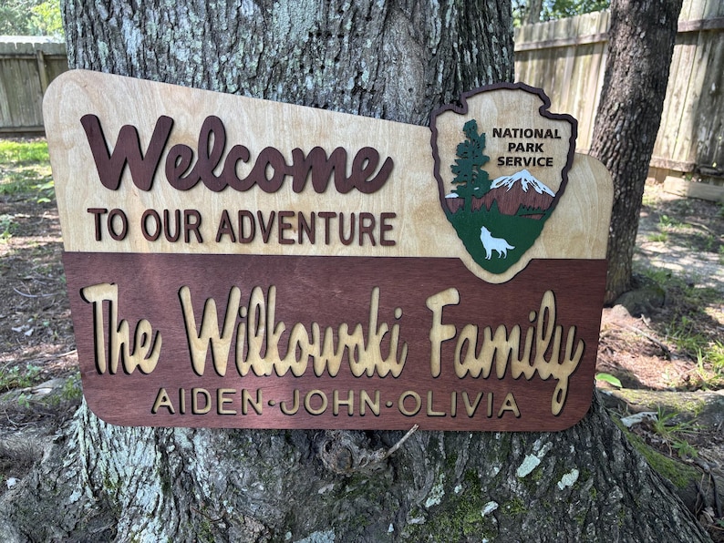 May include: Welcome to our adventure sign with the Wilkowski family name and the names Aiden, John, and Olivia. The sign is made of wood and has a brown and tan color scheme. It features a National Park Service logo with a wolf and a mountain.