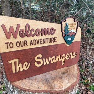 May include: A wooden sign with the text "Welcome to our adventure The Swangers" and the National Park Service logo. The sign is made of two different types of wood, one light and one dark.