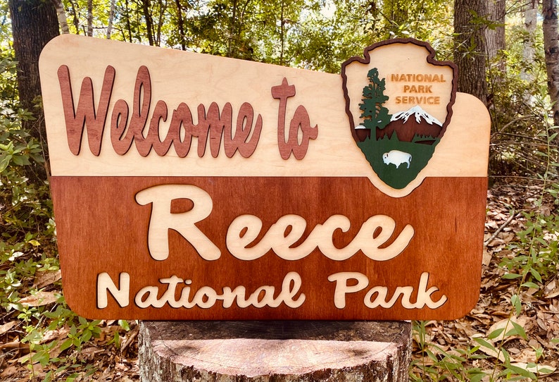May include: Welcome to Reece National Park sign with a brown and beige wood background. The sign has a National Park Service logo with a green background and a white buffalo. The sign is sitting on a tree stump.