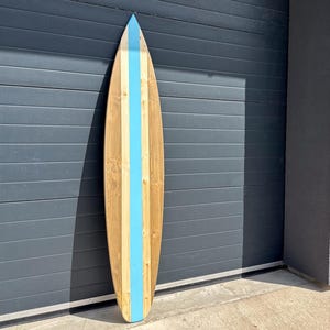 Op de afbeelding: Een houten surfboard met een verticale lichtblauwe streep in het midden. Het board is gemaakt van natuurlijk lichtgekleurde houten planken met zichtbare nerven, met een puntige neus en een gladde, langwerpige vorm.