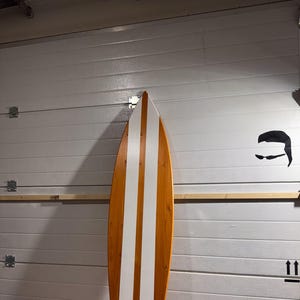 May include: A white and brown surfboard with two white stripes down the centre. The surfboard is leaning against a white wall.