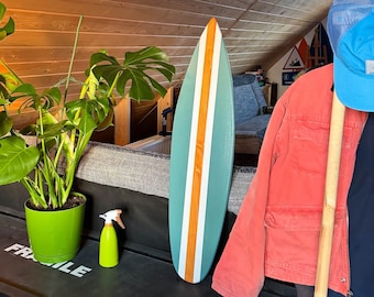 Op de afbeelding: Een turquoise en oranje surfplank leunt tegen een zwart rek met een groene plant in een pot, een spuitfles en een rood jack dat aan een houten rek hangt.