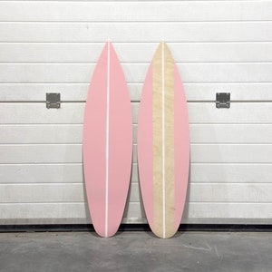 May include: Two decorative surfboards, one pink with a white stripe and one with a natural wood finish and a white stripe. The surfboards are leaning against a white wall.