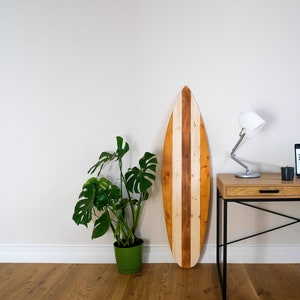 May include: A wooden surfboard with brown and white stripes hangs on a white wall. A desk with a laptop, lamp, and plant is in the foreground.