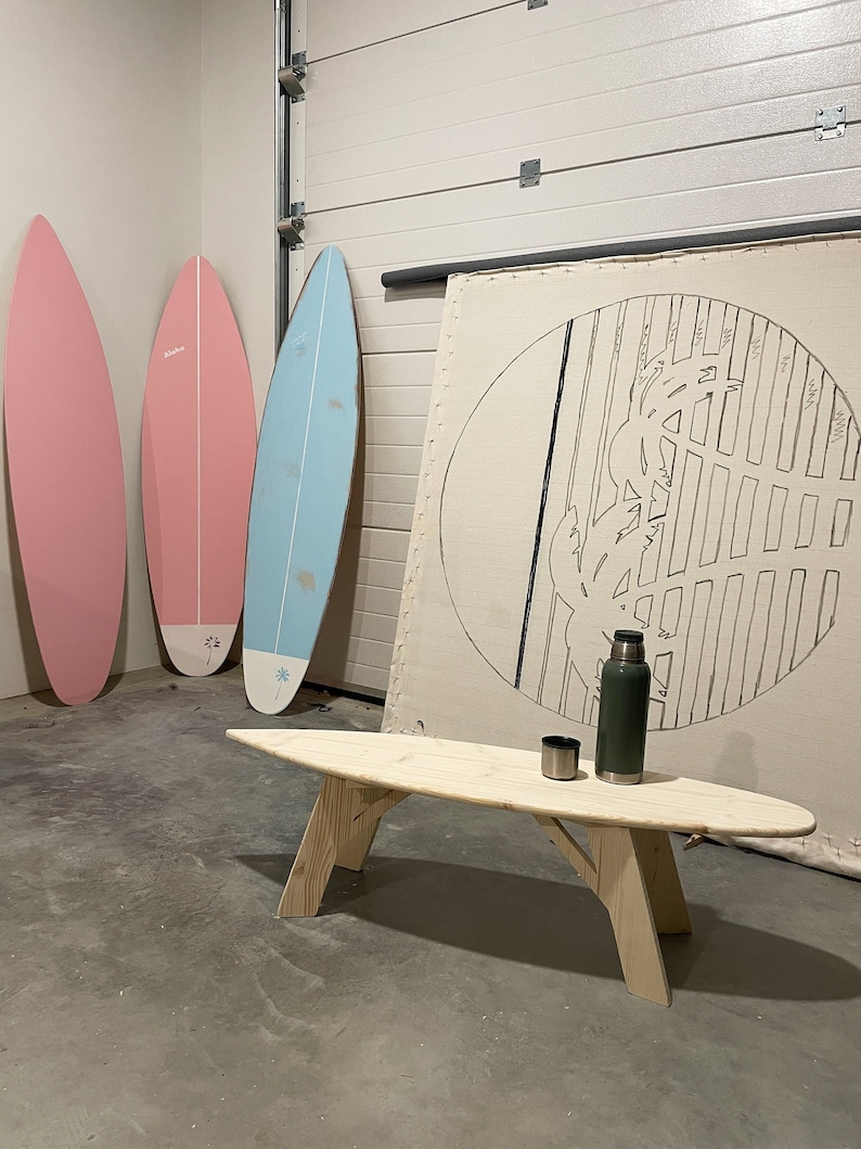 May include: A wooden surfboard coffee table with a light wood finish. The table is sitting in front of a white wall with three surfboards leaning against it. The surfboards are pink, blue, and white. A green flask and a silver cup are on the table.