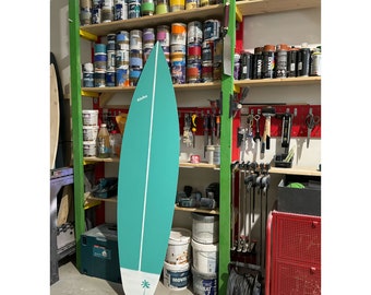 May include: A teal surfboard with a white palm tree design and the word "Alaska" printed on it. The surfboard is leaning against a wall in a workshop.