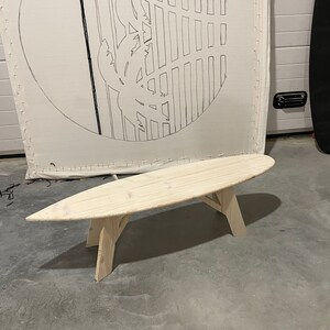 May include: A wooden surfboard-shaped bench with a natural finish. The bench has three legs and is made of light-coloured wood.