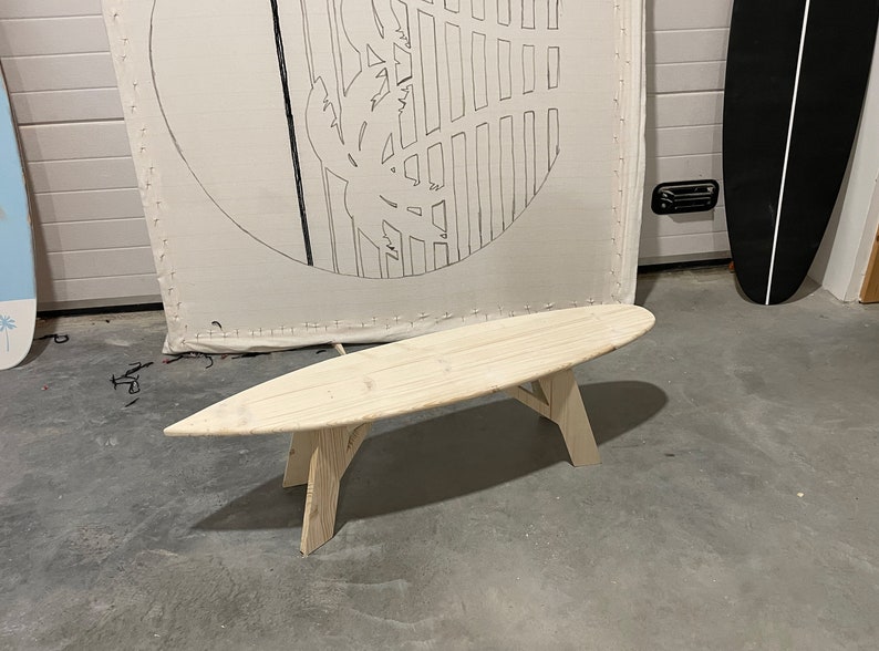 May include: A wooden surfboard-shaped bench with a natural finish. The bench has three legs and is made of light-coloured wood.