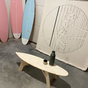 May include: A wooden surfboard coffee table with a green flask and a cup on top. The table is in a garage with three surfboards leaning against the wall.