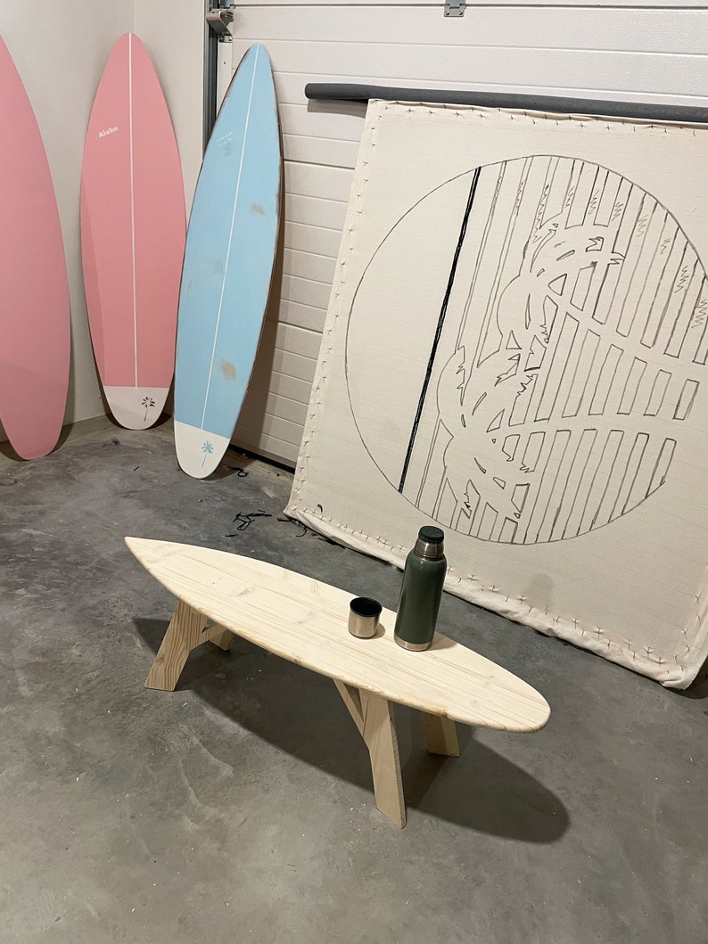 May include: A wooden surfboard coffee table with a green flask and a cup on top. The table is in a garage with three surfboards leaning against the wall.