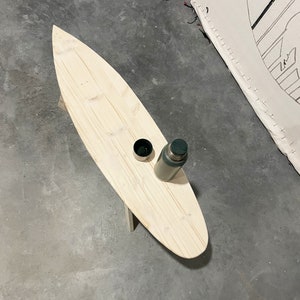 May include: A wooden surfboard-shaped coffee table with a green flask and a small cup on top. The table is sitting on a concrete floor.