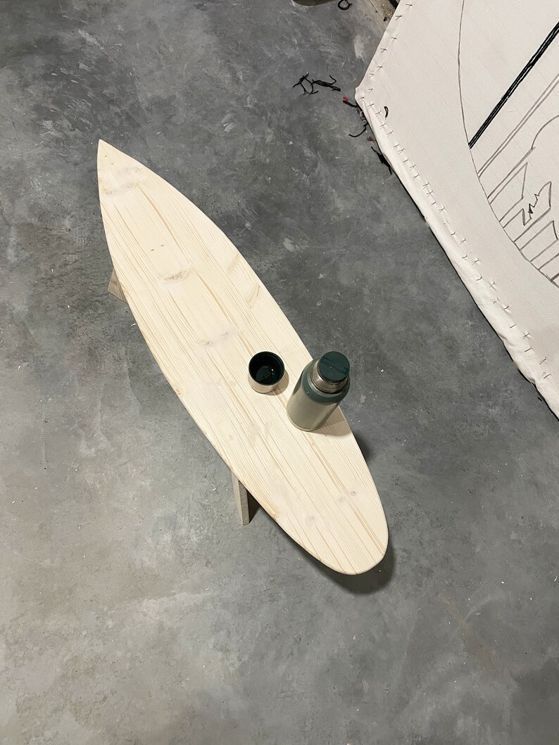 May include: A wooden surfboard-shaped coffee table with a green flask and a small cup on top. The table is sitting on a concrete floor.