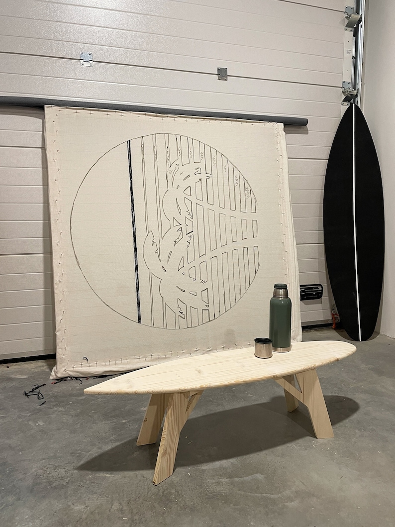 May include: A wooden surfboard coffee table with a light wood finish. The table is in a room with a white canvas with a black circle design and a black surfboard leaning against the wall.