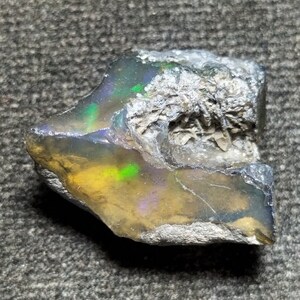 Pode incluir: Um opala bruto, cinza escuro e iridescente com um brilho verde e amarelo. O opala tem uma superfície áspera e natural e é uma pedra bruta não polida.