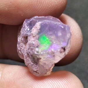 Pode incluir: Um pequeno opala roxo áspero com flashes verdes e iridescentes. O opala está em estado bruto.