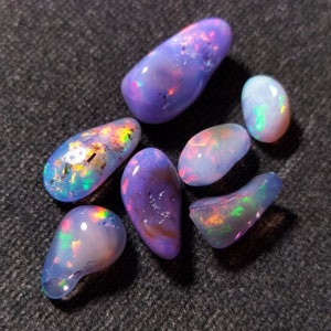 Pode incluir: Sete pedras de opala iridescentes em vários tons de roxo, azul e rosa. As pedras são lisas e têm um brilho perolado.