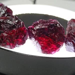 Puede incluir: Primer plano de varias piedras preciosas de granate rojo oscuro en bruto. Las gemas tienen una estructura cristalina y se muestran sobre una superficie blanca con un borde negro. Las piedras están iluminadas desde abajo, resaltando su rico color.