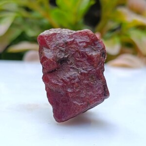 Könnte beinhalten: Ein rauer, ungeschliffener Rubin-Edelstein. Der Rubin hat eine tiefe, satte rote Farbe mit einer strukturierten Oberfläche. Der Edelstein wird vor einem verschwommenen Hintergrund aus grünem und gelbem Laub gezeigt.