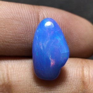 Pode incluir: Um cabochão de opala azul com reflexos iridescentes de cor. A opala tem a forma de uma lágrima e uma superfície lisa e polida.