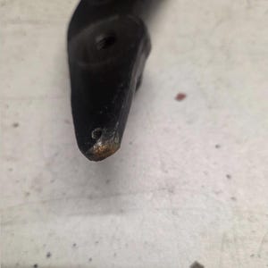 K&ouml;nnte beinhalten: Nahaufnahme eines schwarzen Metallobjekts mit gebogener Form. Das Objekt hat ein Loch und etwas Rost an der Spitze. Der Hintergrund ist eine hellfarbige Oberfl&auml;che.