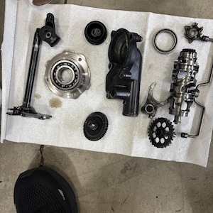 May include: A collection of dismantled mechanical components, including gears, bearings, and a housing, laid out on a white paper towel. The parts are mainly black and silver, possibly from a vehicle or machine.