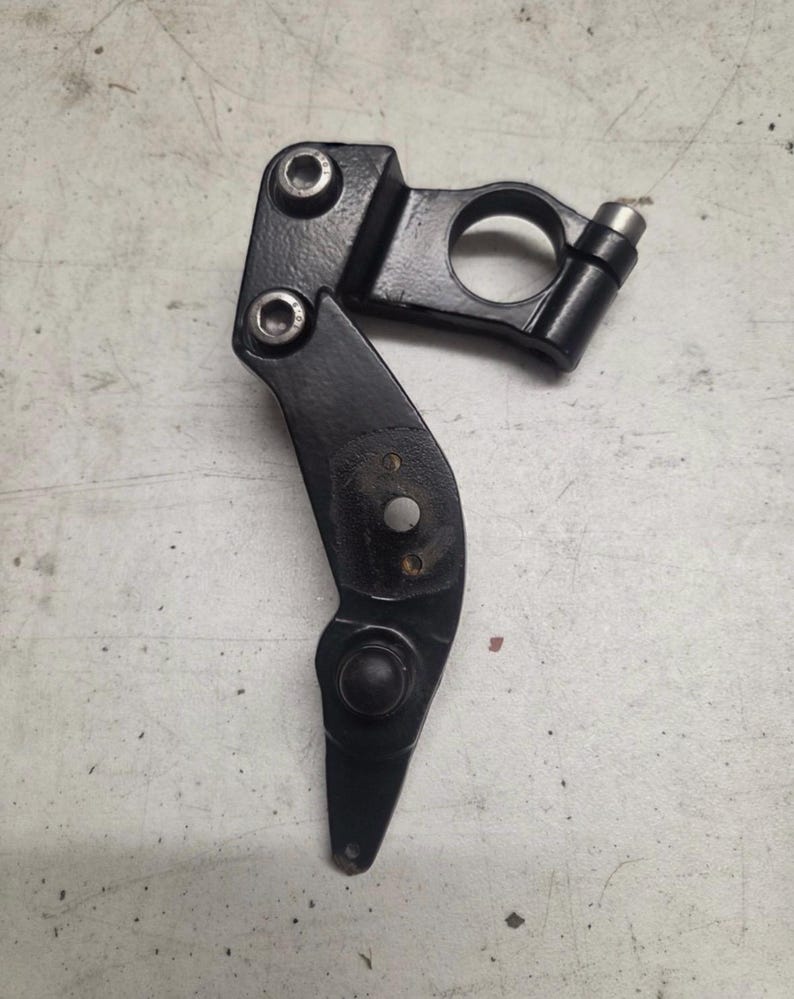 K&ouml;nnte beinhalten: Eine schwarze Metallkomponente mit gebogenem, scharnierartigem Design. Es verf&uuml;gt &uuml;ber eine kreisf&ouml;rmige &Ouml;ffnung, zwei Schraubenl&ouml;cher und einen kleinen zylindrischen Vorsprung. Die Komponente ist wahrscheinlich Teil einer gr&ouml;&szlig;eren mechanischen Baugruppe.