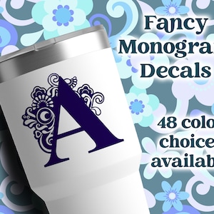 May include: A white stainless steel tumbler featuring a dark purple letter A monogram decal with intricate floral scrollwork. The text on the image reads Fancy Monogram Decals and 48 color choices available. The tumbler has a metallic silver lid.
