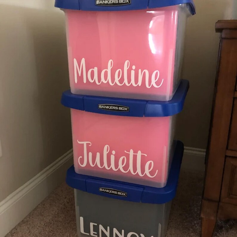 a stack of three storage bins with personalized names on them. The first bin is pink and has the name "Madeeline" written on it. The second bin is also pink and has the name "Juliette" written on it. The third bin is gray and has the name "Lennon" written on it.