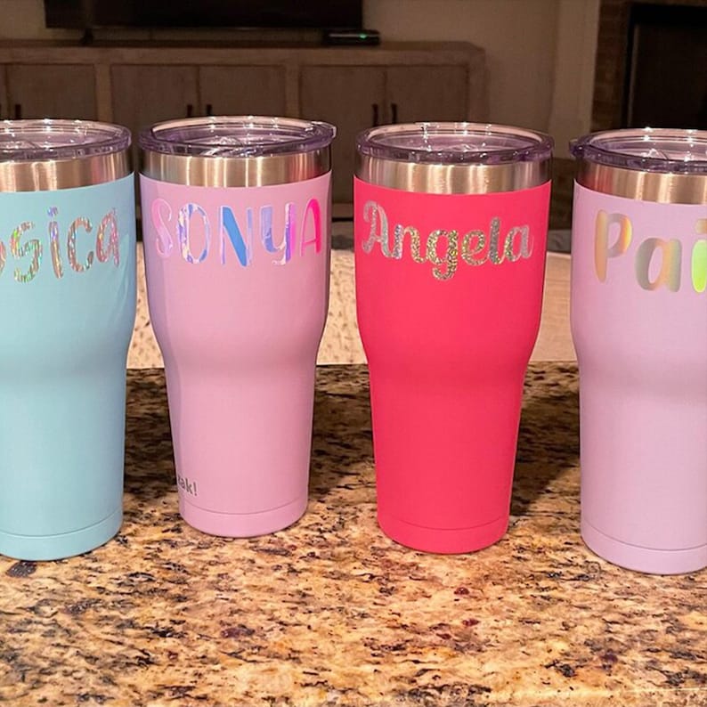 A row of four colorful personalized tumblers with names printed on them, sitting on a granite countertop.