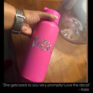A person's hand holding a pink water bottle with a personalized decal, and a glass container in the background.