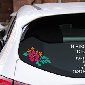 May include: A white car's rear window features a colorful hibiscus decal. The decal includes a pink flower with orange accents and green leaves. Text on the window reads "Hibiscus Decal for Tumblers, Mugs, Cars, Coolers & Lots More."