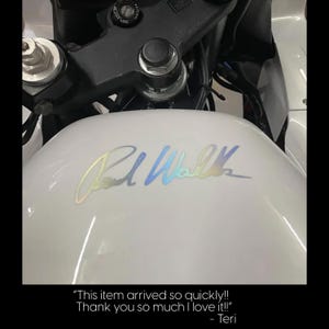 a close-up view of a white motorcycle, focusing on the gas tank with a signature or logo on it.