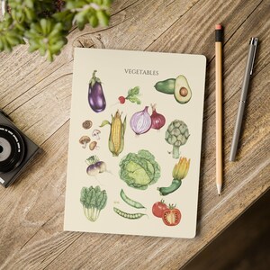 May include: A notebook with a beige cover displaying watercolour illustrations of vegetables such as aubergine, avocado, and tomatoes. The word "VEGETABLES" is printed at the top. A pencil and pen are on the right.