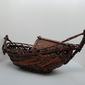 May include: A woven brown basket shaped like a boat with a long handle. The basket is made of natural materials and has a rustic appearance.