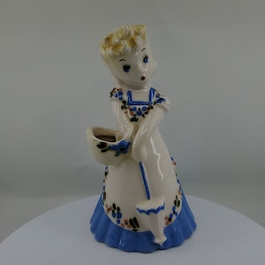 May include: A ceramic figurine of a girl wearing a white dress with blue trim and floral accents. She is holding a blue watering can and a basket with flowers.