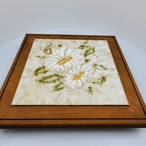 May include: A framed ceramic tile with a white background and a floral design featuring two white flowers with yellow centers and green leaves.