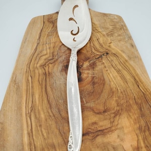 May include: Silver cake server with a decorative cut-out design on the blade and a floral pattern on the handle. The server is resting on a wooden cutting board.