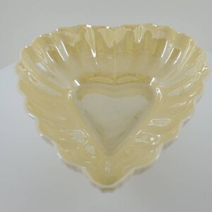 May include: A heart-shaped ceramic dish with a scalloped edge. The dish is a pale yellow color and has a glossy finish.