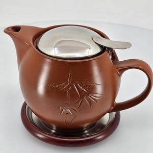 Può includere: Teiera in ceramica marrone con coperchio e infusore in acciaio inossidabile, appoggiata su un sottobicchiere in legno con base in acciaio inossidabile. La teiera presenta un disegno di bambù inciso.