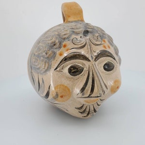 May include: A ceramic pitcher shaped like a face with a brown handle. The face is painted with black and brown lines and has orange accents. The pitcher has a whimsical and folk art style.