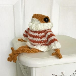 May include: A handmade, knitted frog figurine wearing a white and orange striped sweater and a white turtleneck. The frog has large black eyes and orange legs. The frog is sitting on a white surface.